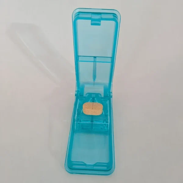 Un diviseur-pilule bleu ouvert avec un comprimé beige positionné dans le support en V prêt à être coupé.