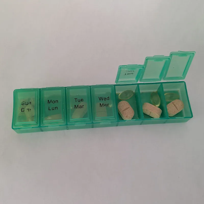 The green pillbox with several lids open, showing various pills and capsules organized by day.