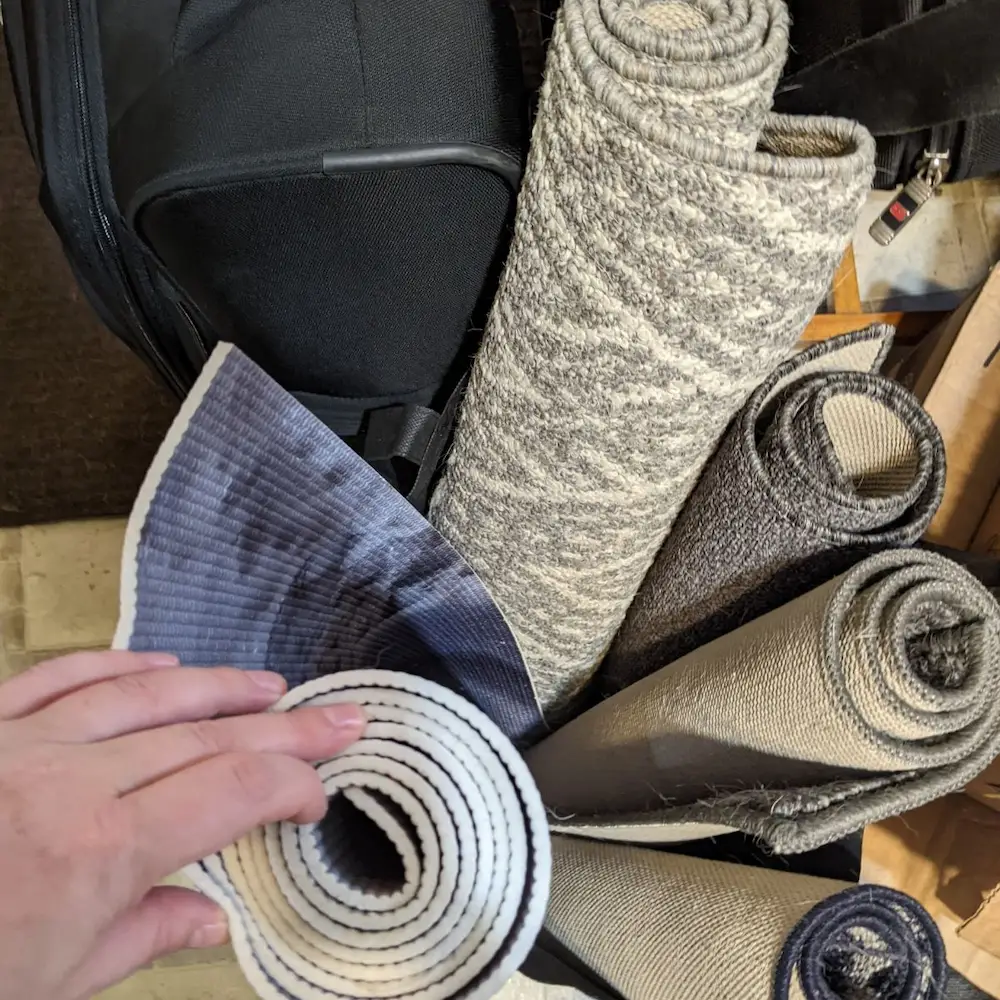A collection of rolled-up non-slip yoga mats and rugs packed into a travel bag, prepared as a mobile kit to provide traction for an older dog at a sitter's home.